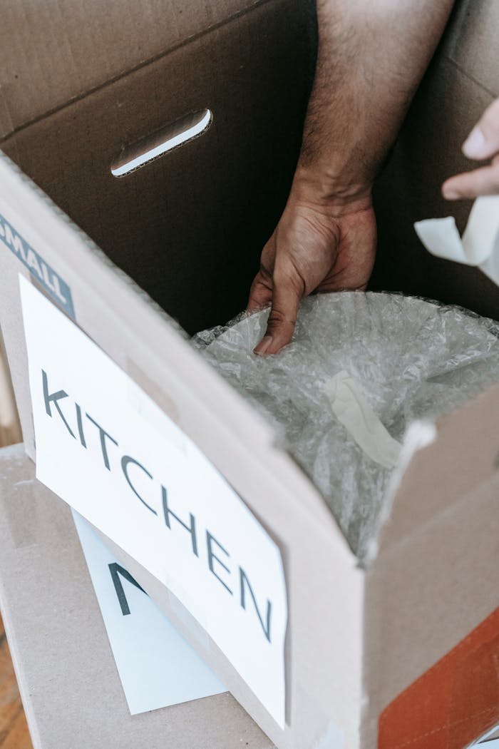 hero-img A close-up view of wrapping kitchen items for moving in a labeled cardboard box indoors.