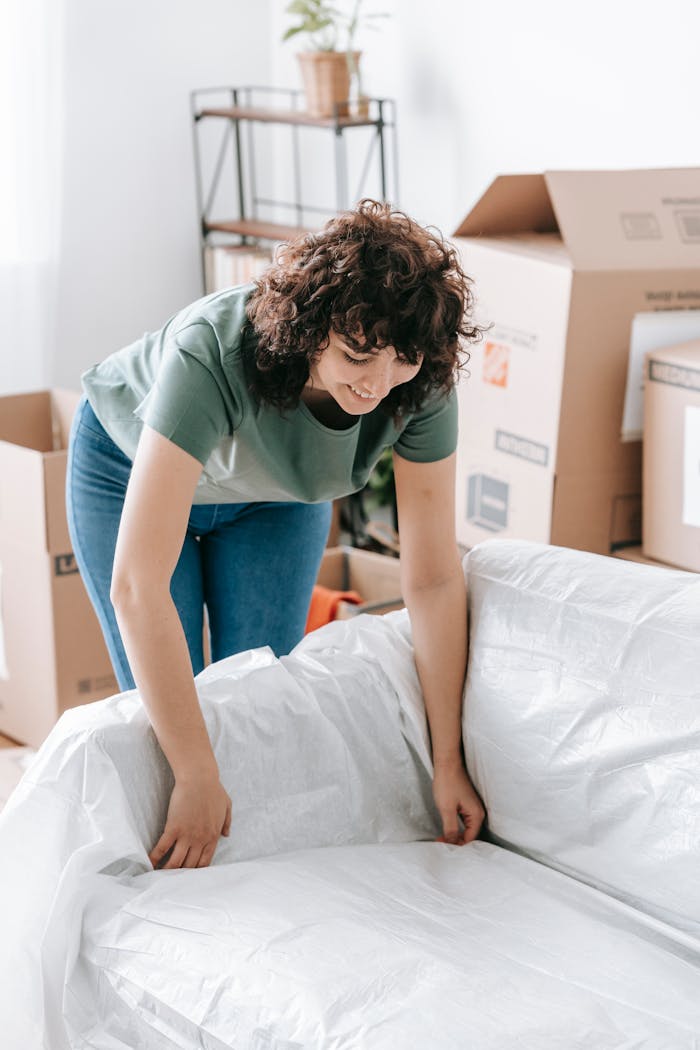 about-01 Woman wrapping a sofa with plastic for moving in a bright living room.
